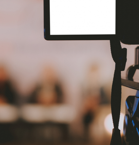 Billedet viser et kamera og en række mennesker, der taler ved en pressekonference